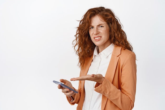 Frustrated redhead saleswoman pointing at mobile phone grimacing and staring confused standing in suit against white background