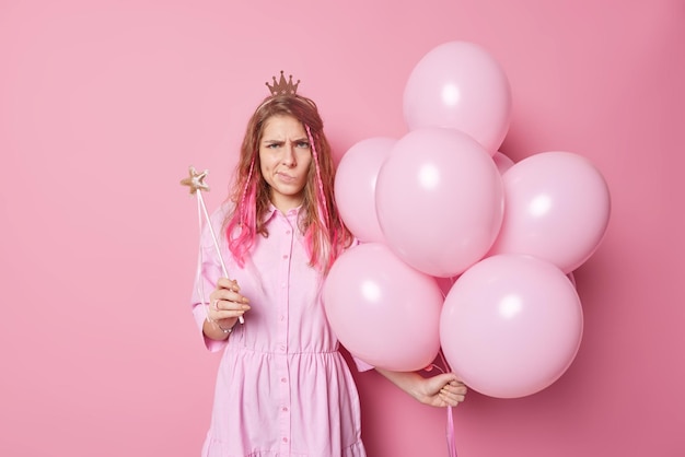 Frustrated unhappy long haired young woman holds bunch of inflated balloons and magic wand being angry with someone wears dress and crown poses against pink background doesnt like something