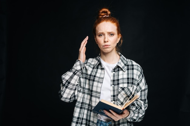 Frustrated upset young woman college student holding book and looking at camera on isolated black background Pretty redhead lady model emotionally showing facial expressions in studio copy space