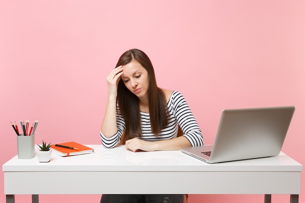 Frustrated woman in desperation leaning on hand looking down sit, work at white desk with contemporary pc laptop