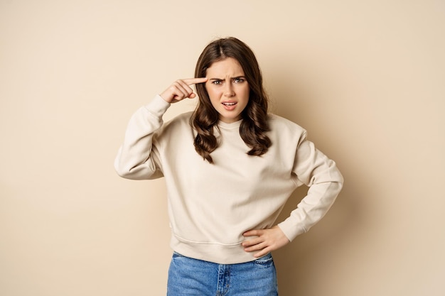 Frustrated woman roll finger near head and complaining insulting someone standing over beige background