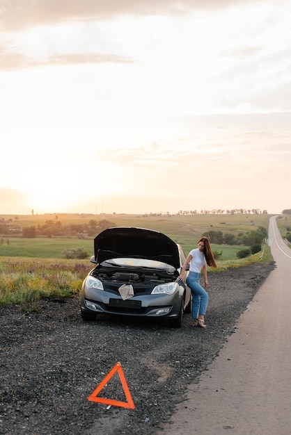 A frustrated young girl stands near a brokendown car in the middle of the highway during sunset Breakdown and repair of the car Waiting for help Car service Car breakdown on road