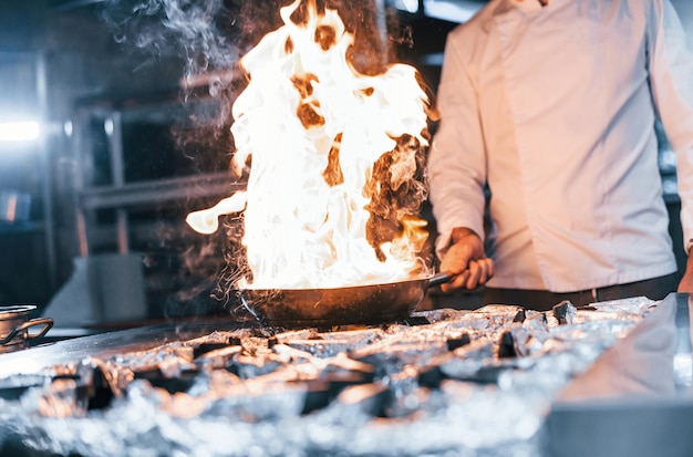 Frying pan is on fire Chef in white uniform cooking food at kitchen Busy day at work