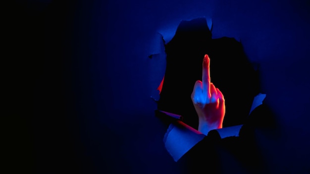 Photo fuck you provocative gesture red blue neon light female hand showing middle finger obscene sign inside breakthrough paper hole isolated on dark night copy space banner