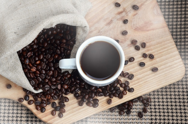 A full bag of brown coffee beans and a white cup of hot coffee lies on a wooden surface