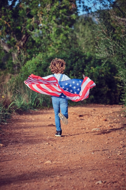 Full body back view of anonymous active boy running with colorful USA flag on rural path in countryside with green trees