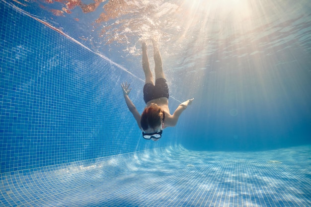 Full body of barefoot boy in goggles and shorts swimming in clean pool under sunshine