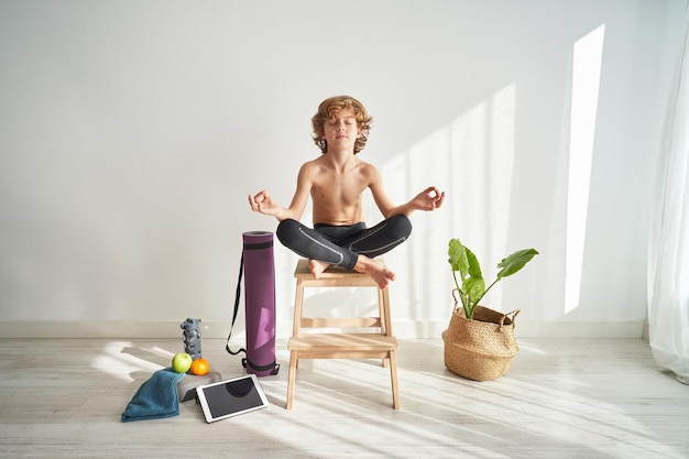 Full body of barefoot preteen boy sitting with crossed legs on wooden stool and relaxing in Padmasana yoga pose