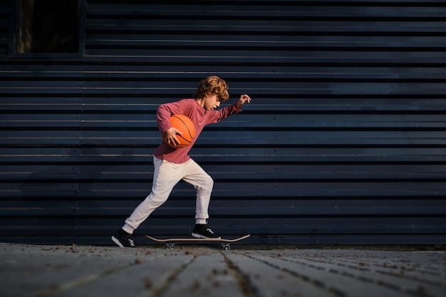 Full body boy with basketball ball riding skateboard on pavement near dark blue wall