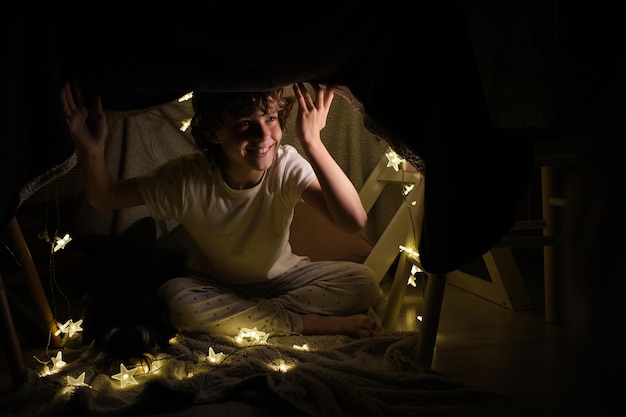 Full body of cheerful boy looking away while sitting under blanket construction with glowing garland in dark room at home