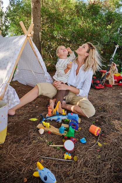 Full body of cheerful mother and son in casual outfits sitting on grass near wigwam and playing with various colorful toys while looking up in park