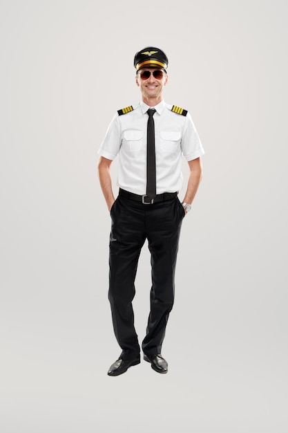 Full body of confident adult male aviator in uniform and sunglasses keeping hands in pockets and smiling friendly, while looking at camera against gray background