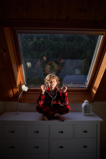 Full body of cute little boy in sleepwear sitting on chest of drawers with crossed legs in Lotus pose and with hands in mudra gesture at mansard window in evening