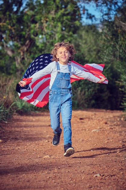 Full body of delighted boy looking at camera while running on road with national waving American flag on summer day