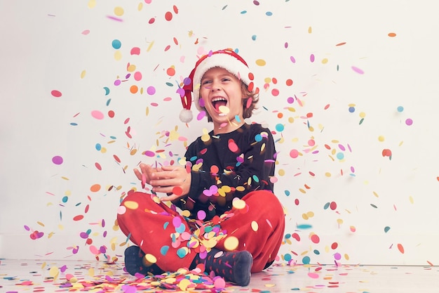 Full body of delighted teen boy in Santa hat looking up while sitting under bright falling confetti on white background during Christmas celebration