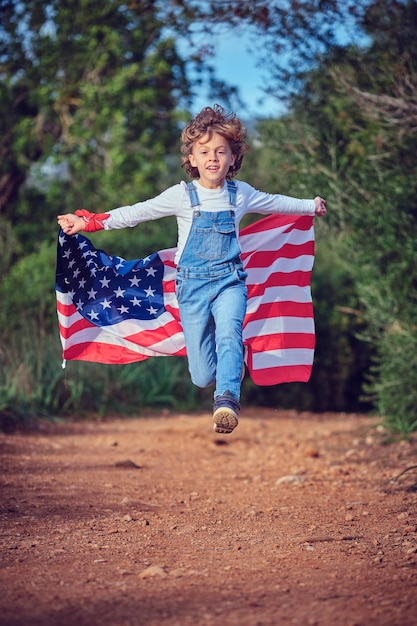 Full body of energetic boy with colorful waving American flag running fast on rural road in countryside on summer day