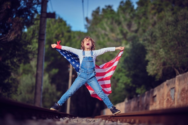 Full body of expressive kid holding national USA flag while standing on rails against green trees in daylight