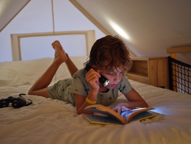 Full body of focused barefoot boy in nightwear reading book with glowing flashlight while lying on comfortable bed on mansard