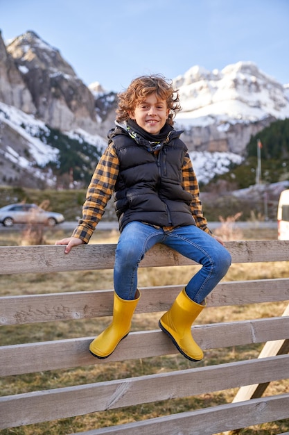 Full body of happy boy with curly hair in warm clothes sitting on wooden fence while smiling and looking at camera in nature