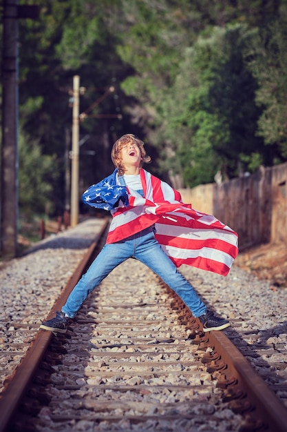 Full body of happy preteen boy rolled in USA flag rejoicing over national holiday and standing on rails of railroad