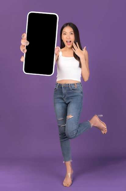 Full body length photo portrait of beautiful Asian young woman Excited surprised girl showing big smart phone with blank screen white screen isolated on purple background Mock Up Image