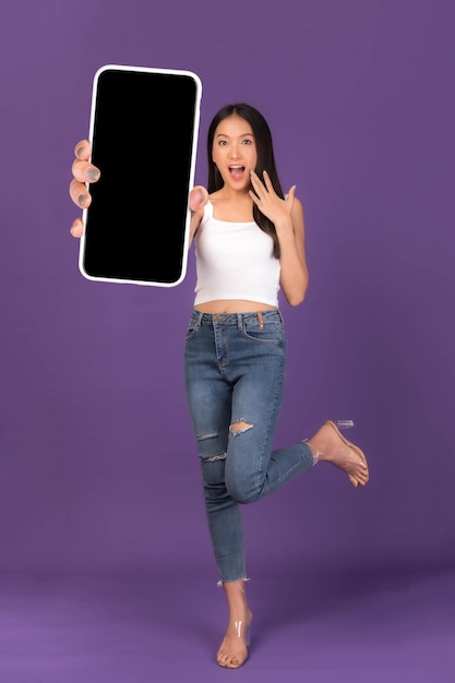 Full body length photo portrait of beautiful Asian young woman Excited surprised girl showing big smart phone with blank screen white screen isolated on purple background Mock Up Image