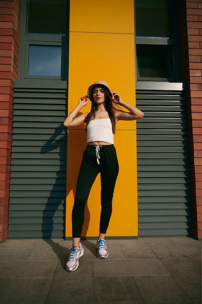 Photo full body of modern millennial fit female wearing skinny top with leggings and hat standing near urban building