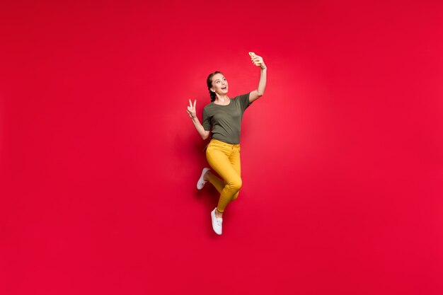 Full body photo of crazy funny lady jumping high making selfies showing v-sign symbol cheerful mood wear casual yellow pants green t-shirt isolated red color background