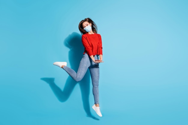 Full body photo of lady jump high up good mood wear mask knitted red pullover sneakers jeans isolated blue color background