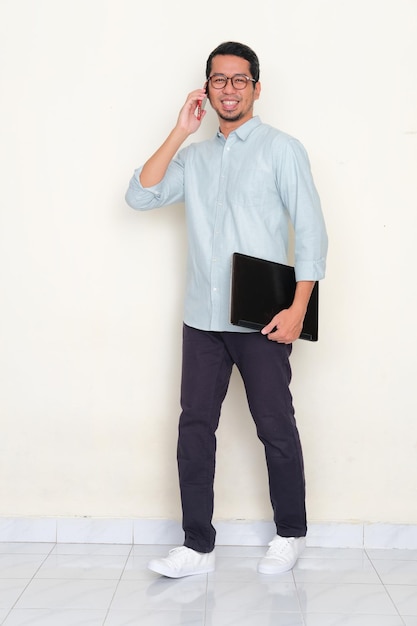 Full body portrait of Asian man walking while answering a phone call and holding laptop