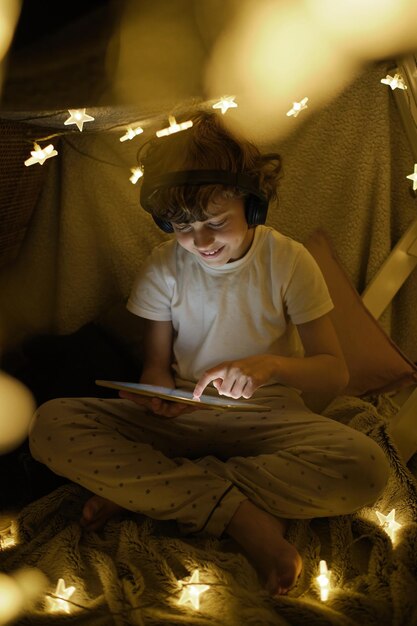 Full body of positive boy in headphones enjoying video on table sitting with crossed legs in blanket tent with glowing garlands