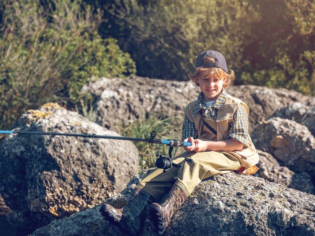 Full body of positive boy in vest sitting on rocky coast with fishing rod while catching fish on summer day