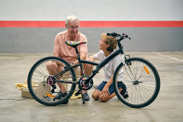 Full body of positive senior man repairing bicycle with cheerful grandson laughing with opened moth