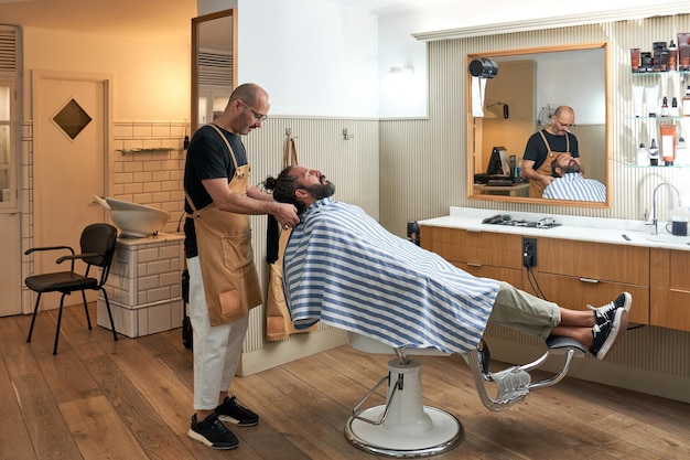 Full body of professional male coiffeur in beige apron grooming adult bearded man in cozy barbershop