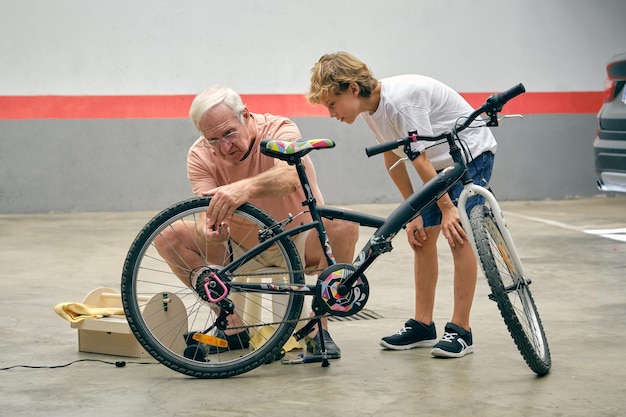 Full body of serious grandfather inflating tires of bicycle near grandson looking at wheel with attention