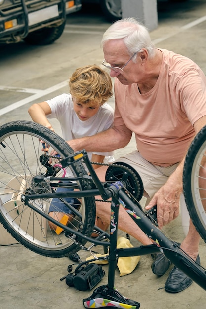 Full body of serious grandfather inflating tires of bicycle near grandson looking at wheel with attention