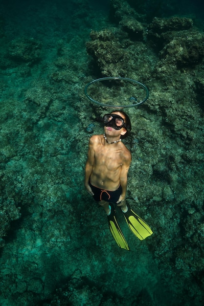 Full body of shirtless boy in snorkeling mask and flippers swimming through bubble ring in deep sea during vacation