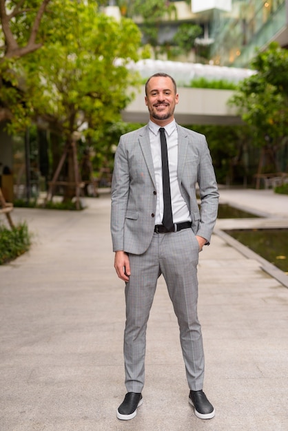 Full body shot of happy Hispanic bald bearded businessman smiling with nature in the city