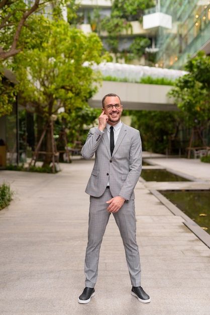 Full body shot of happy Hispanic bald bearded businessman wearing eyeglasses with nature in the city