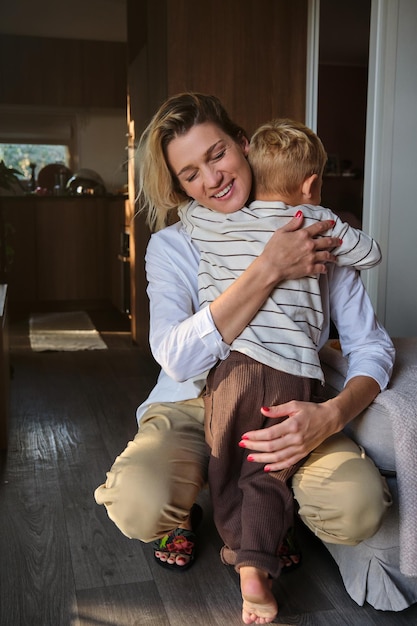 Full body of smiling woman in white blouse hugging adorable barefoot kid while leaning on sofa in dark apartment with daylight