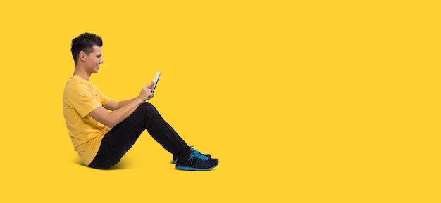 Full body Teenage boy portrait sitting using tablet computer isolated on yellow background