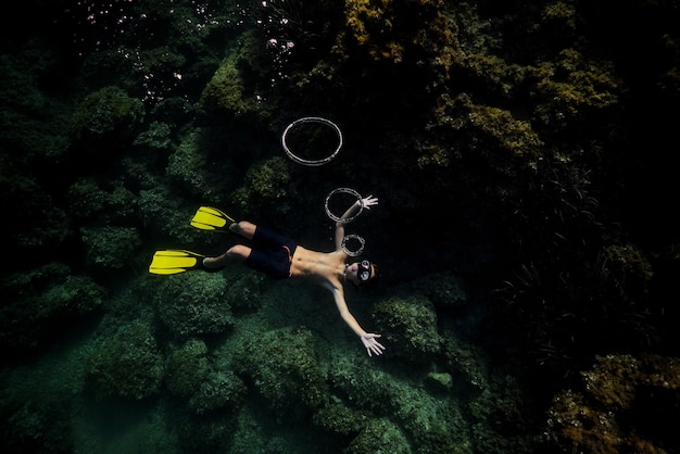 Full body top view of anonymous male diver in wetsuit and flippers swimming under deep dark seawater near coral reefs and blowing ring bubbles