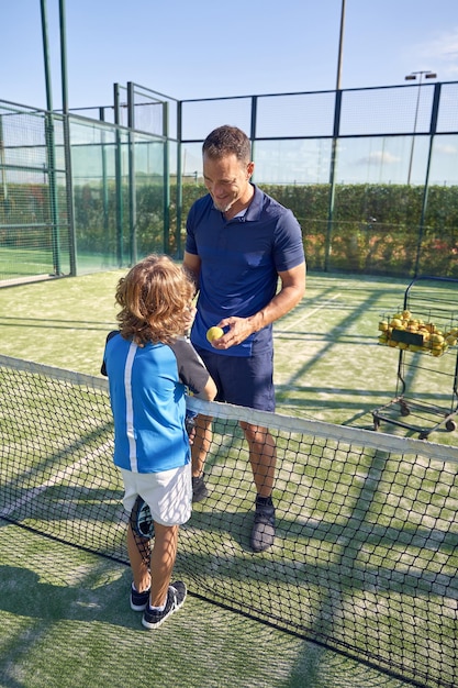 Full body of trainer speaking with boy in activewear while standing near net during padel tennis training on court in summer day