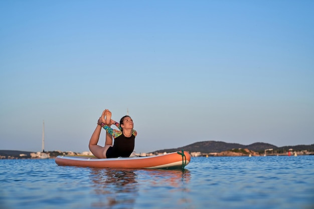 Full body of young flexible woman in swimwear practicing yoga in Dhanurasana on SUP board surrounded by rippling sea against picturesque scenery