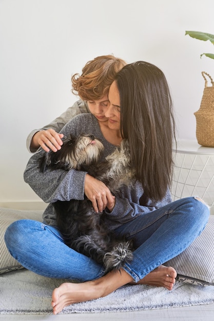 Full body of young long haired woman in jeans and curly haired boy embracing fluffy puppy and looking at pet while sitting on floor in modern living room