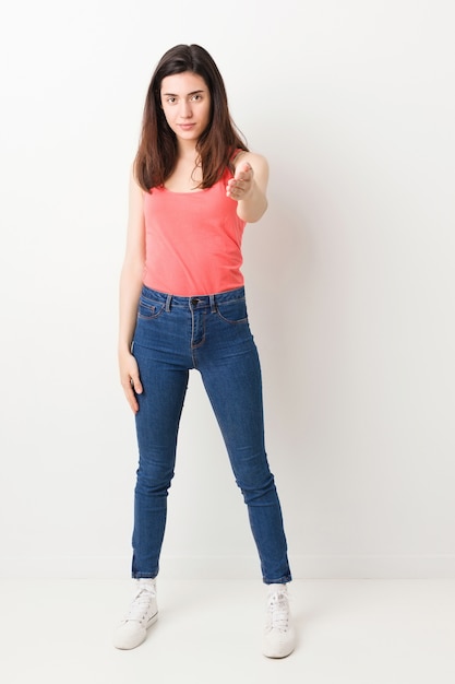 Full body young woman on white wall stretching hand at camera in greeting gesture.