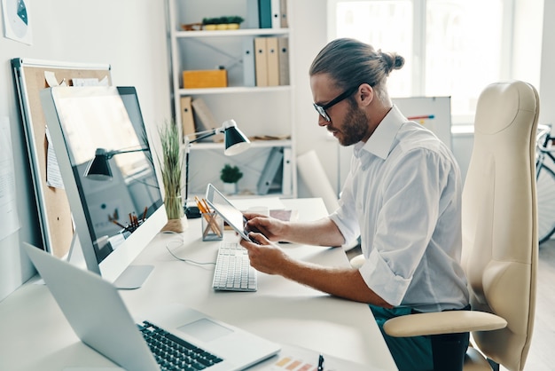 Full concentration. Good looking young man in shirt using digital tablet while sitting in the office