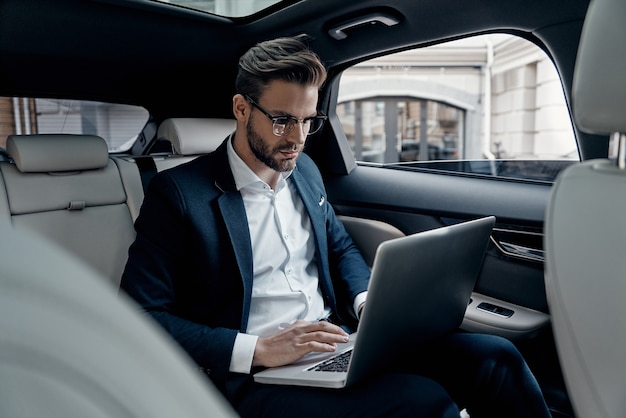 Full concentration at work. Confident young man in full suit working using laptop while sitting in the car