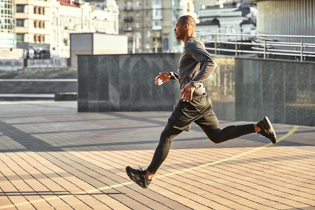 Full of energy Full length portrait of athletic african man running with very fast speed through the sunny city streets. Side view. Healthy lifestyle. Sport and motivation concept. Urban workout