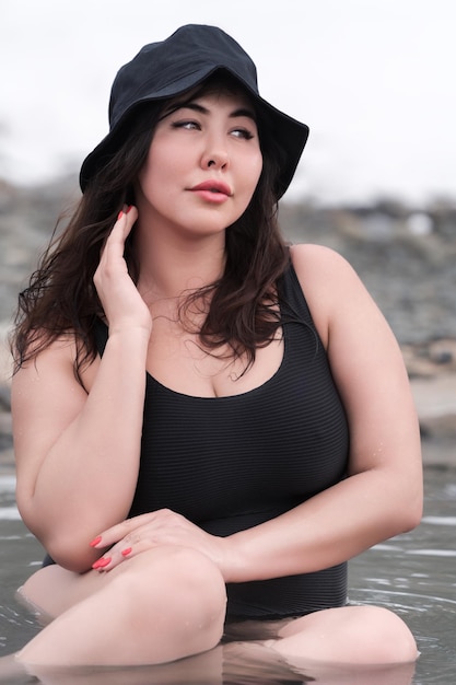 Full figured young model in black bathing swimsuit and panama hat sitting in outdoors pool at spa